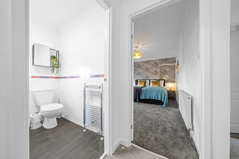 Hallway with open bathroom door showing toilet and towel rack, bedroom visible ahead