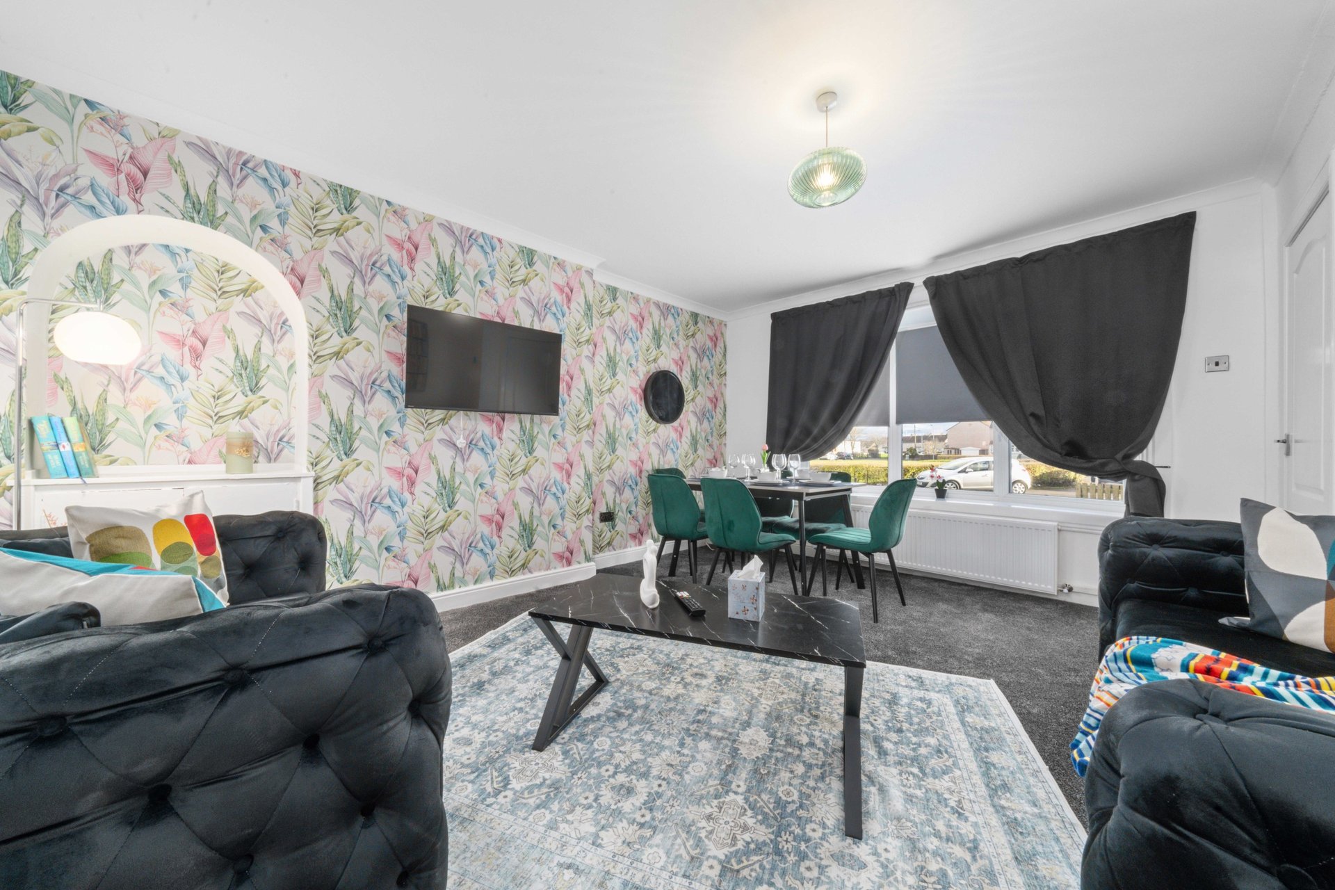 Open-plan living room with floral wallpaper, black furniture, desk area and mounted TV