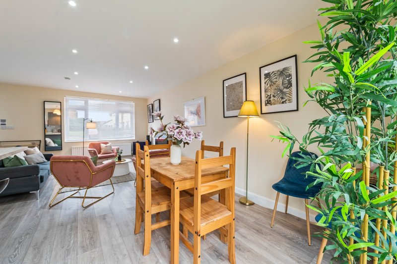 Open-plan living and dining area with wooden dining table, upholstered armchairs, potted plants and framed wall art