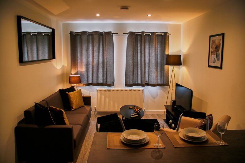 Modern living room with black sofa, dining table set for two, and curtained windows with warm lighting