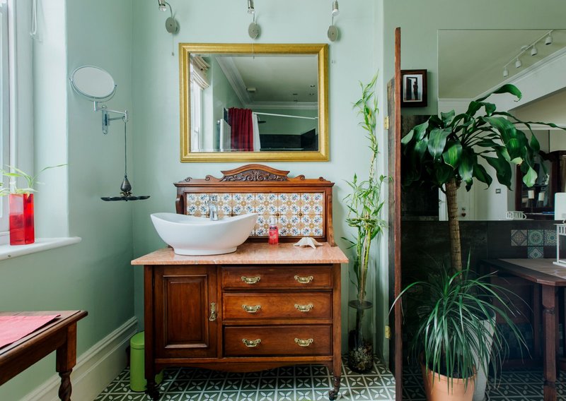 Bathroom with sage green walls, wooden vanity with vessel sink, large gold-framed mirror, potted plants