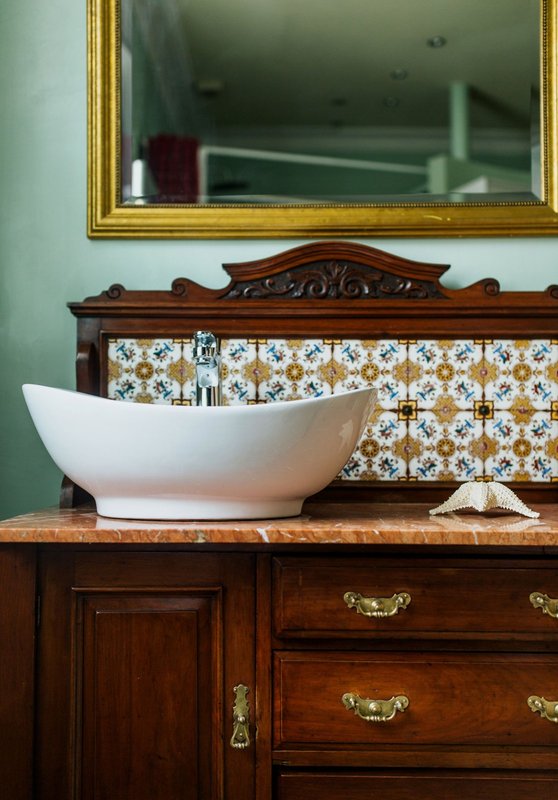 Bathroom vanity with white ceramic basin, wooden cabinetry, decorative tile backsplash and gold-framed mirror