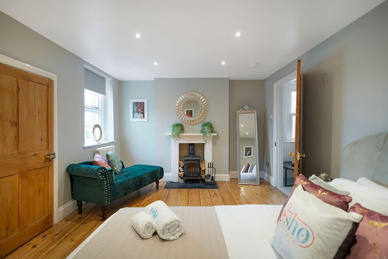Spacious bedroom with wooden flooring, fireplace, green velvet seating, and ensuite door