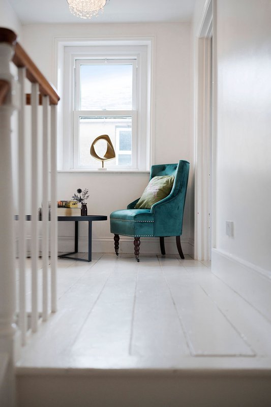 Bright hallway with teal armchair, small side table, window and white wooden flooring