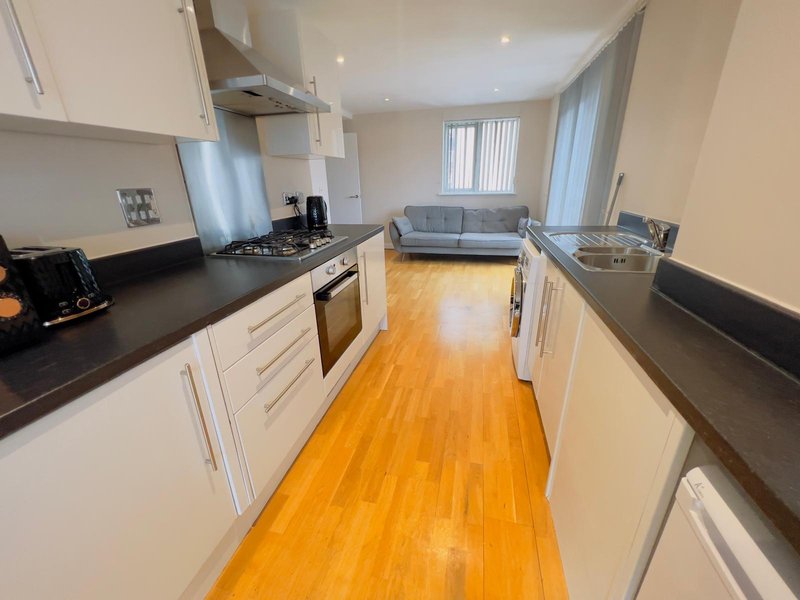 Modern galley kitchen with white cabinetry, black countertop, wooden flooring and sofa visible beyond