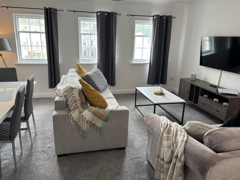 Spacious living room with cream upholstered sofa, TV, multiple windows with black curtains, and neutral décor