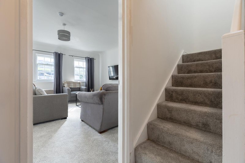 Hallway with carpeted staircase leading upward, doorway to living room with grey armchairs and windows