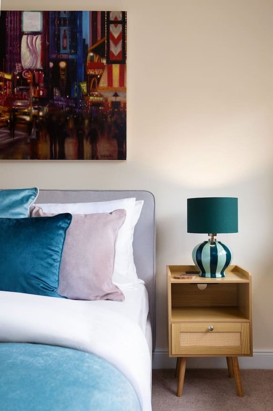 Bedroom with blue upholstered bed frame, white bedding, teal and purple cushions, wooden nightstand with lamp and framed artwork above.