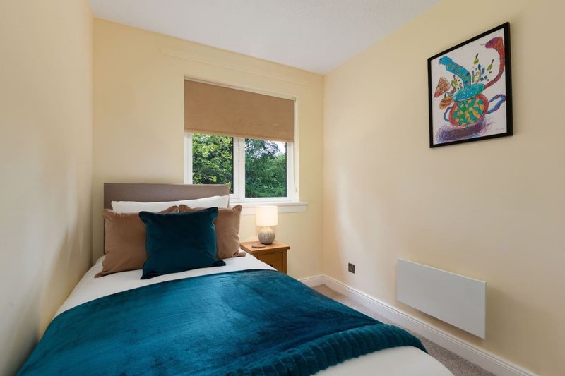 Compact bedroom with single bed, teal bedding, wooden nightstand, and window with blinds
