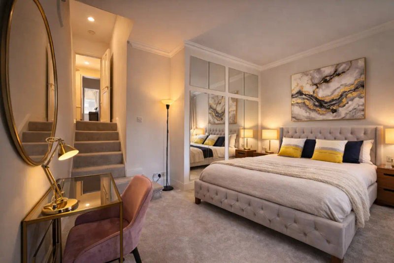 Elegant bedroom with upholstered bed, gold-framed mirror, stairs, and warm lighting