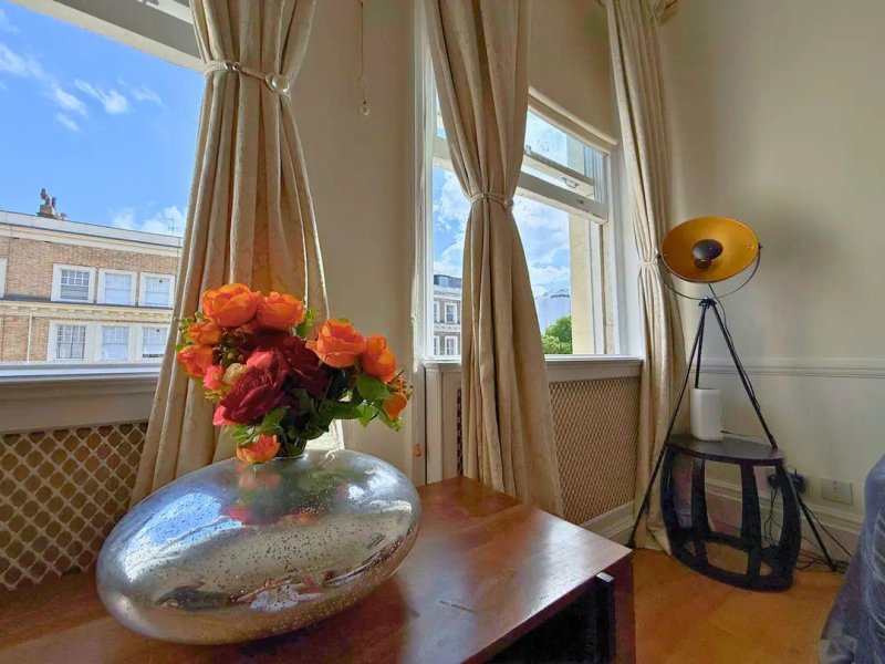 Bright living room with wooden table, orange flowers in vase, floor lamp, and tall windows with city views