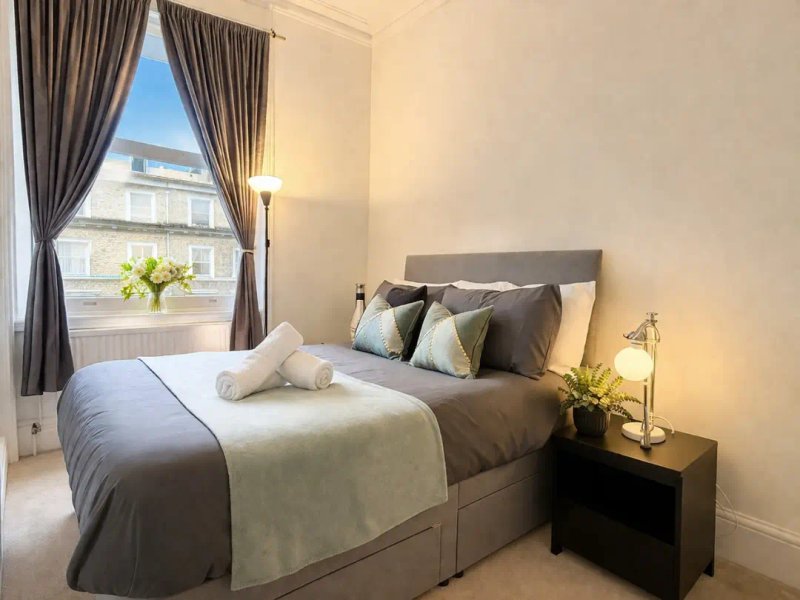 Modern bedroom with double bed, grey headboard, cushions, bedside lamp and window with curtains