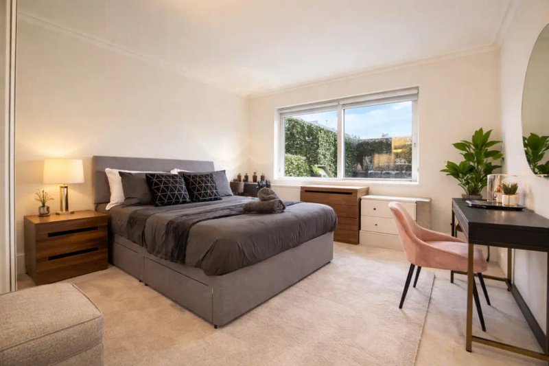 Spacious bedroom with grey bed, wooden nightstand, desk area, and large windows overlooking garden