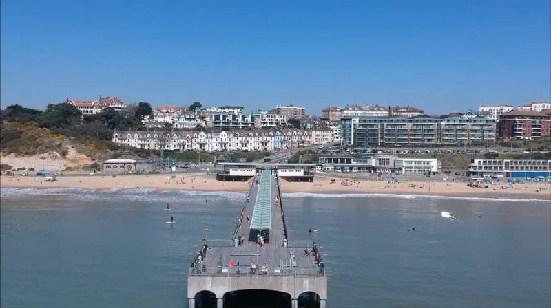 Beach pier extending into calm blue water with seaside town and buildings visible in background