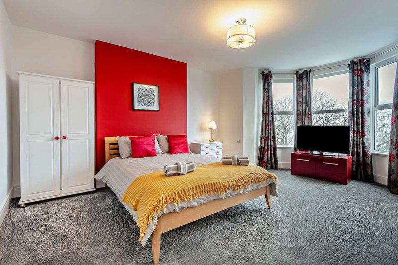 Bedroom with red accent wall, double bed with yellow throw, white wardrobe and TV unit