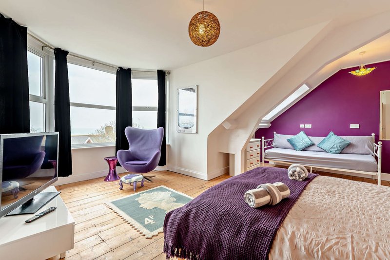 Spacious bedroom with purple accent wall, double bed, armchair, desk with monitor and wooden flooring