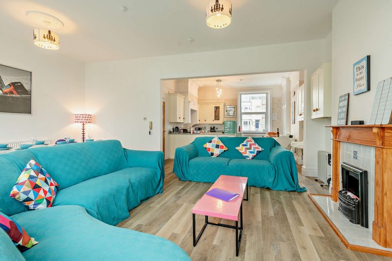 Bright living room with turquoise sofas, colorful cushions, wooden flooring, and open-plan kitchen visible beyond