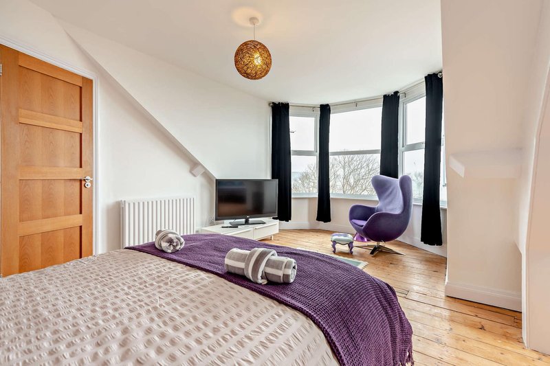 Bright bedroom with double bed, purple throw, wooden floor, window seat and TV