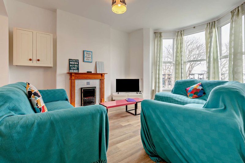 Bright living room with two turquoise sofas, fireplace, TV, and large windows overlooking garden