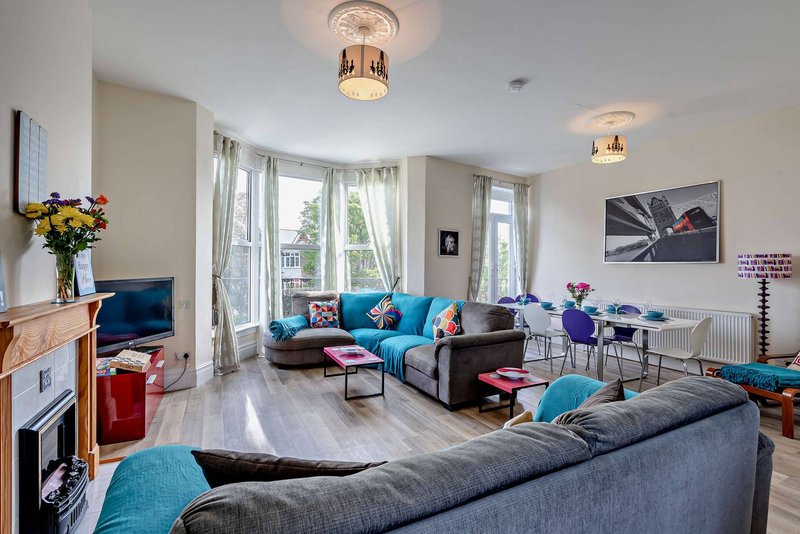 Bright open-plan living room with two grey sofas, turquoise cushions, wooden flooring and large windows
