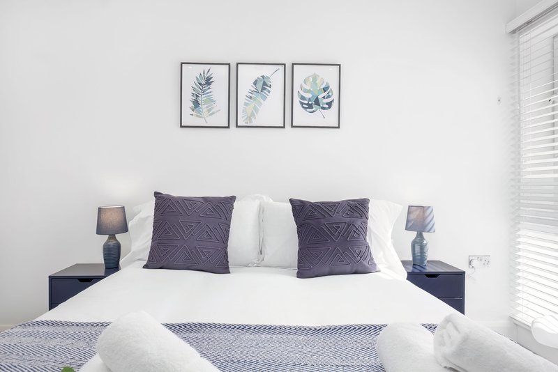 Modern bedroom with white bedding, purple cushions, dark nightstands and botanical wall art