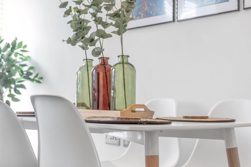 Dining table with white chairs, decorative vases, fresh greenery, and natural light from windows
