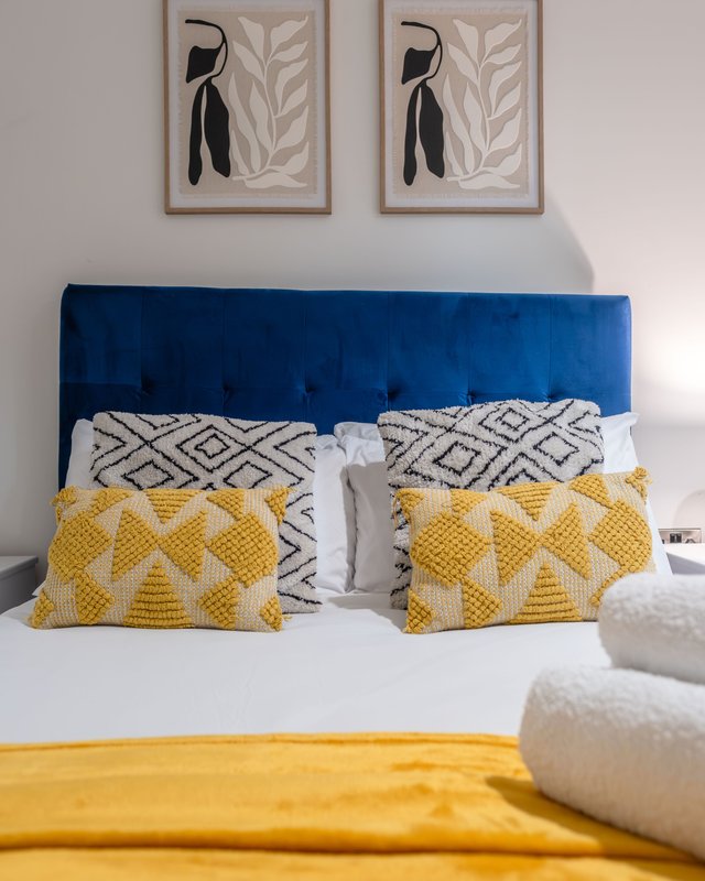 Bedroom with blue upholstered headboard, patterned pillows, yellow bedding, and framed wall art