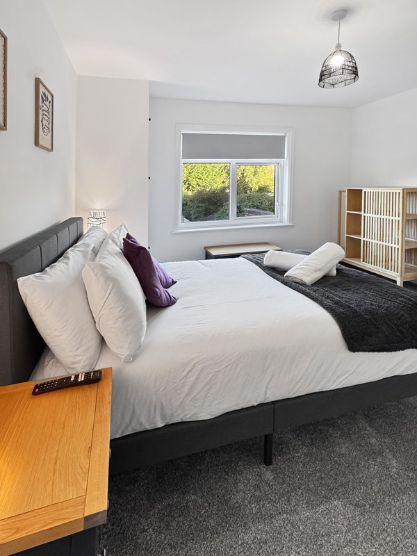 Bright bedroom with double bed, white bedding, purple cushion, wooden nightstand and window.