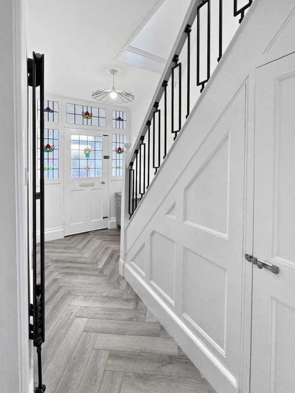 Bright hallway with herringbone wood flooring, white walls, staircase with black railing, and front door with decorative window panels