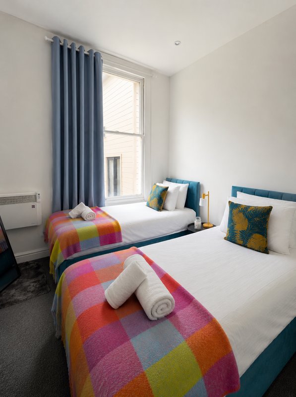 Twin bedroom with white walls, blue curtains, colorful patterned bedding and window view