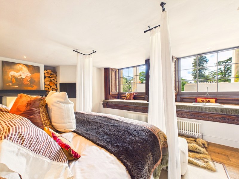 Modern bedroom with four-poster bed, orange and gold cushions, dark bedding, and large windows with views