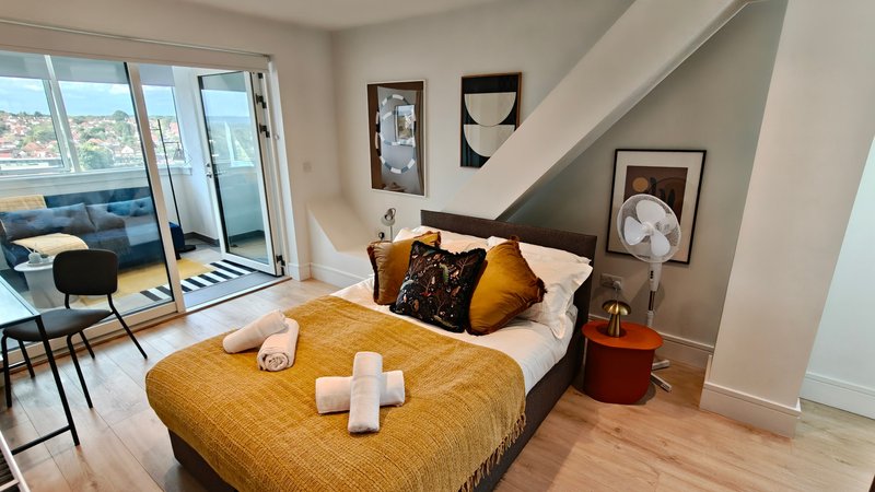 Modern bedroom with double bed, gold bedding, white pillows, framed wall art, and balcony doors with city views