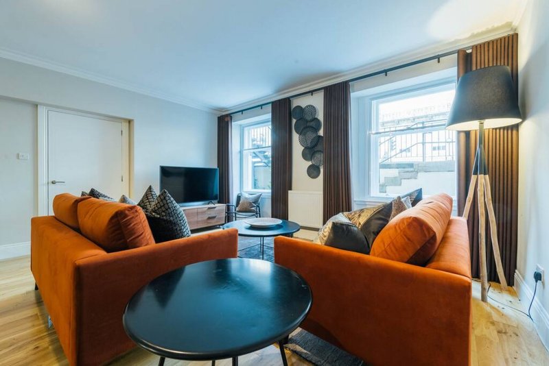 Modern living room with two orange sofas, round black table, TV, and large windows with city views