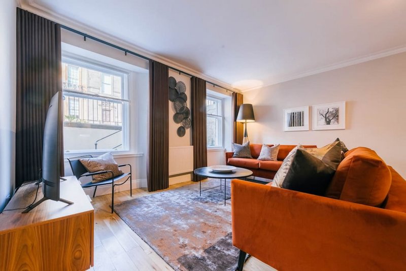 Modern living room with orange sofa, large windows, curtains, wooden furniture and framed wall art