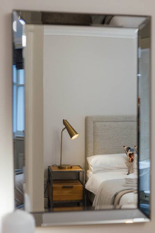 Bedroom reflected in mirror showing bed with headboard, wooden nightstand and brass lamp