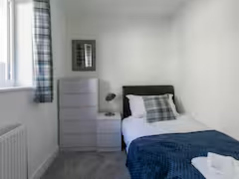 Compact bedroom with single bed, grey chest of drawers, small mirror and checkered curtains
