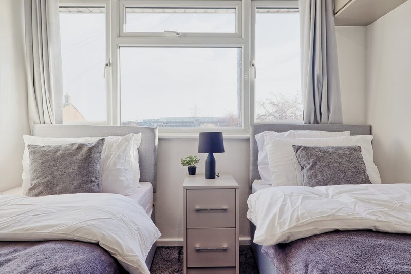 Twin bedroom with two beds, grey headboard, nightstand, lamp, and large window with curtains