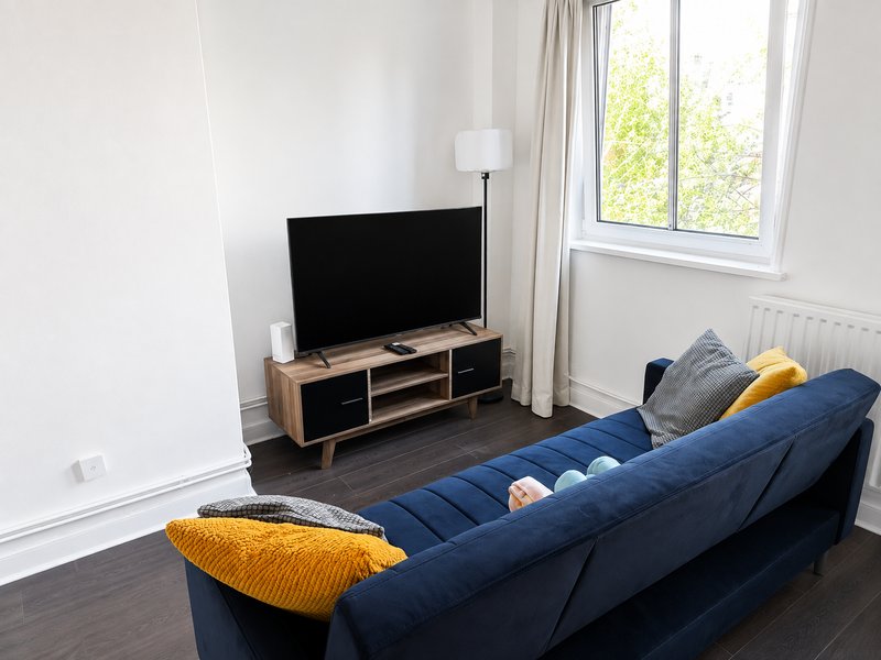 Living room with blue sofa, TV on wooden stand, yellow cushions, and large window with curtains