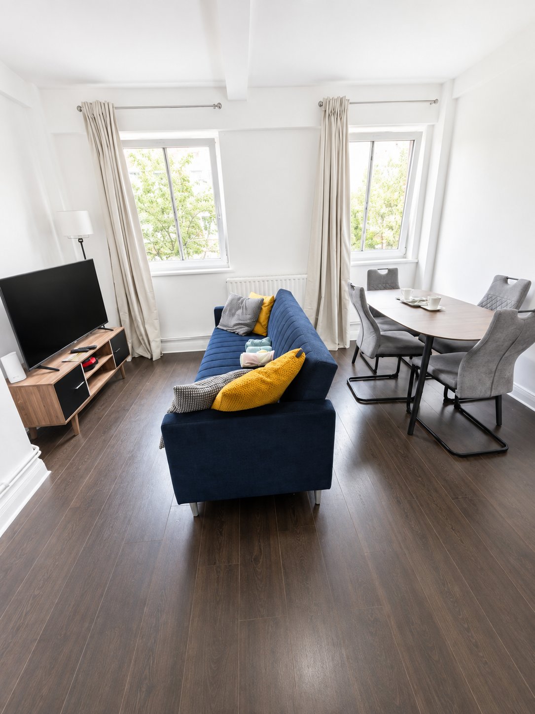 Bright open-plan living room with blue sofa, yellow cushions, TV, dining table and large windows