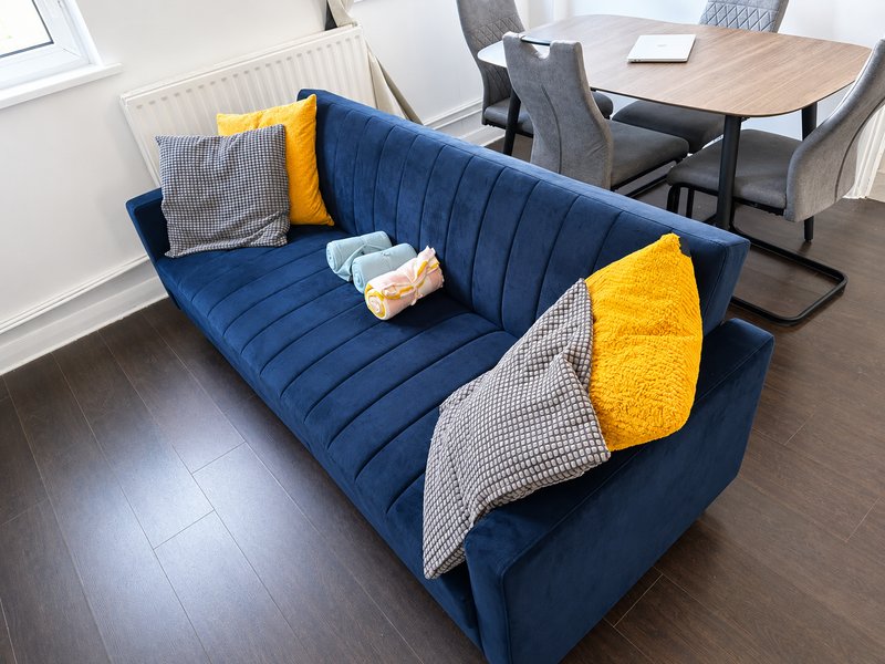 Blue upholstered sofa with yellow and grey cushions, dining table and chairs in background
