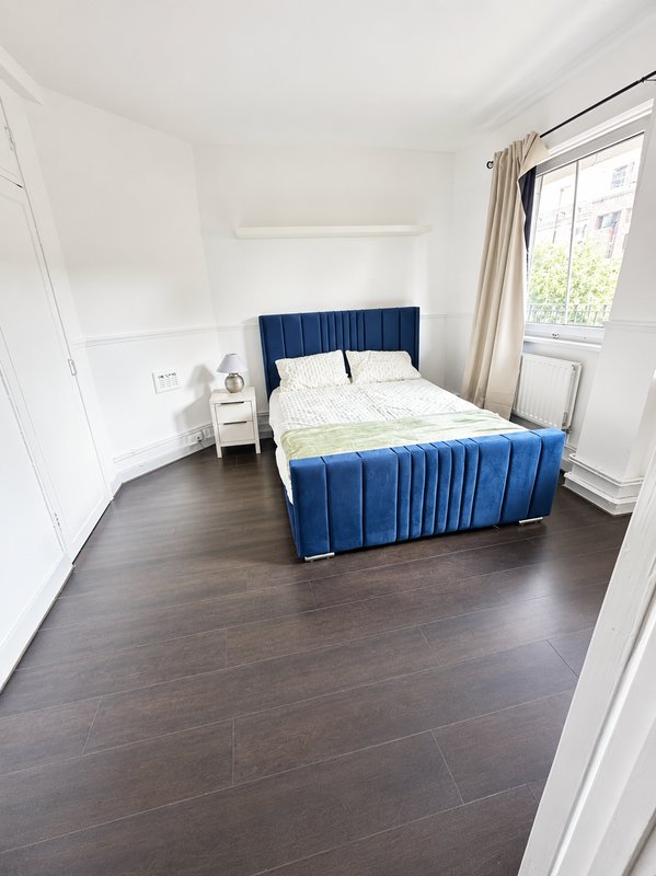 Bright bedroom with blue upholstered bed, dark wood flooring, white walls and window with curtains
