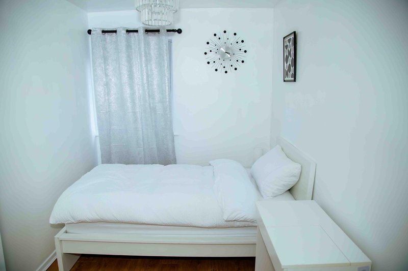 Small bedroom with single bed, white bedding, sheer curtains and wall clock
