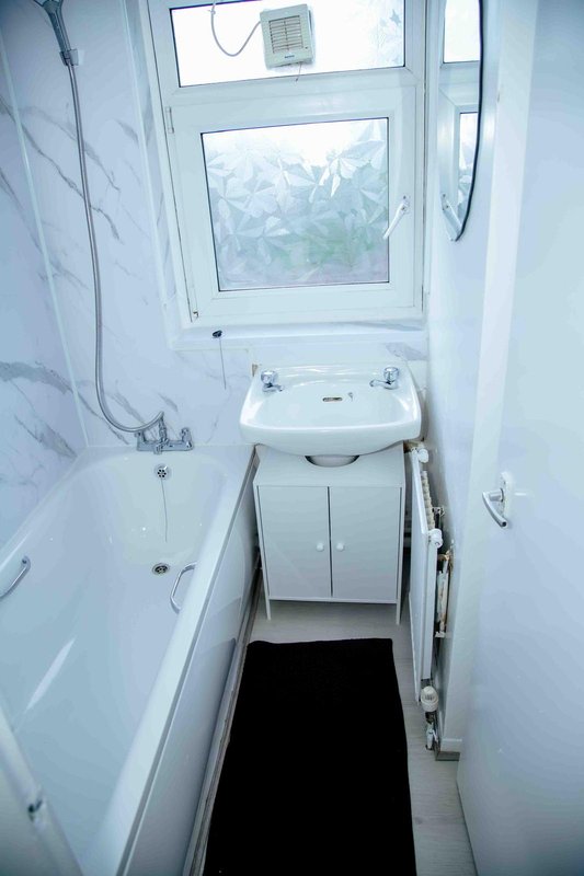 Narrow bathroom with white sink, shower enclosure, window and dark floor