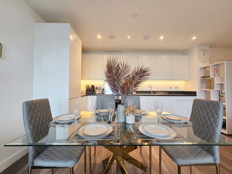 Modern dining room with glass table, grey upholstered chairs, and white kitchen beyond