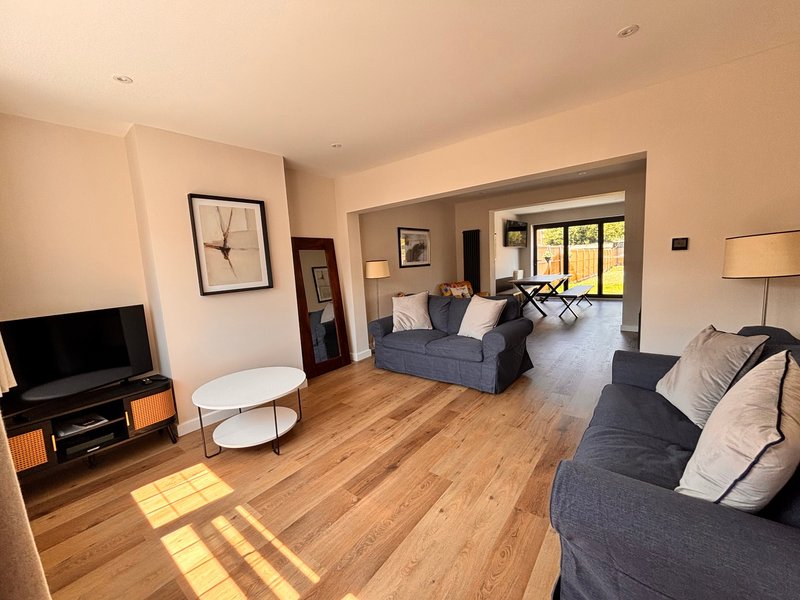Open-plan living area with wooden flooring, blue sofas, TV unit, and bright windows