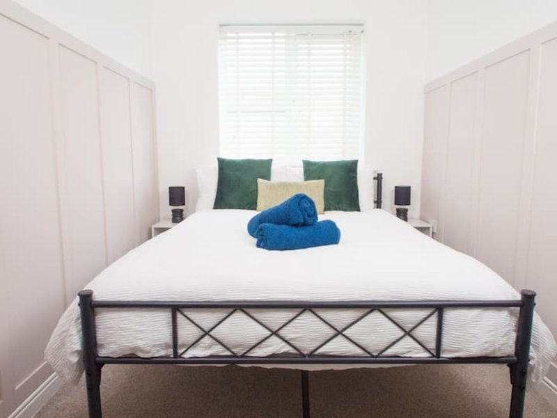 Bright bedroom with metal-frame bed, white bedding, green and cream pillows, blue towel roll