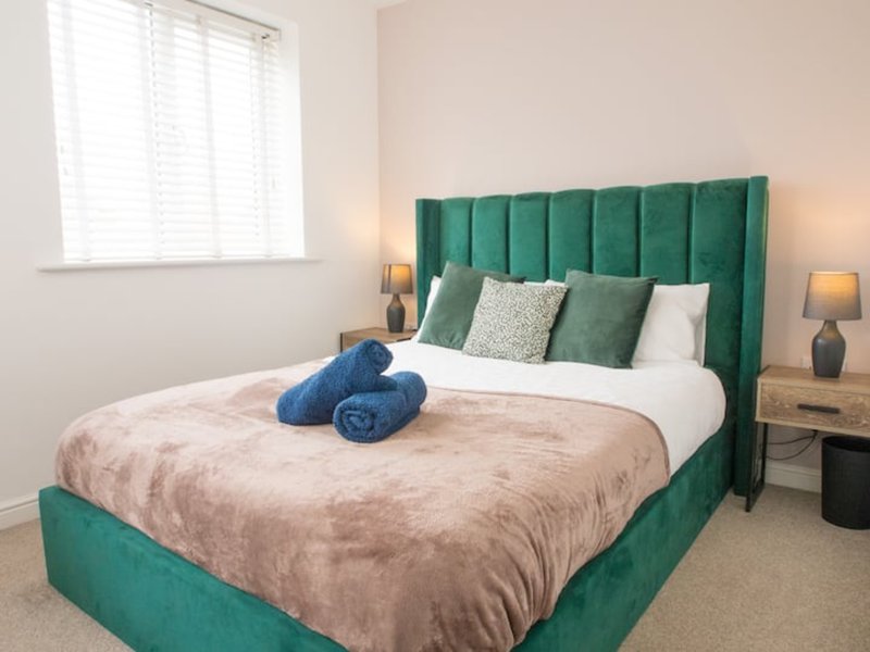 Bedroom with green upholstered bed, white pillows, beige bedding and blue towels