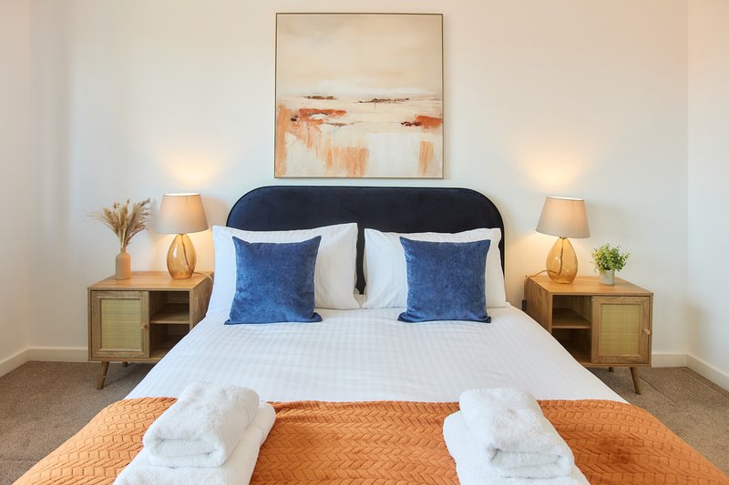 Modern bedroom with dark headboard, white bedding, blue cushions, and matching wooden nightstands with lamps