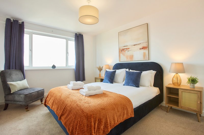 Bright bedroom with double bed, orange throw, armchair, wooden nightstand and wall art