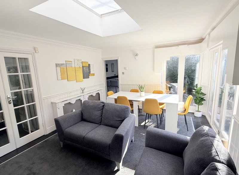Bright open-plan living and dining area with dark sofas, wooden dining table, yellow chairs and garden doors